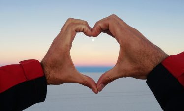 Ein mit den Händen gebildetes Herz umfasst den Vollmond der über das Salar de Uyuni untergeht